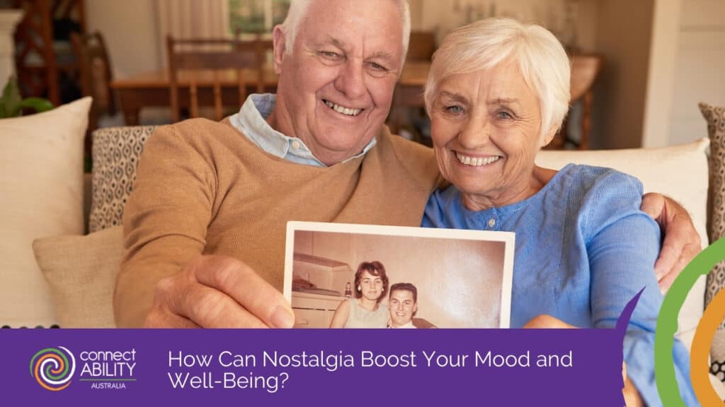 A smiling elderly couple sits together on a couch holding up an old photograph of themselves as a young couple