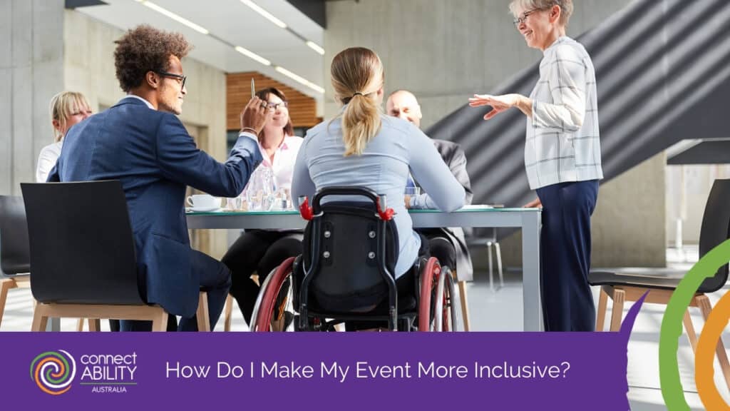 A woman in a wheelchair sits at a meeting table with colleagues while a woman standing at the head of the table speaks