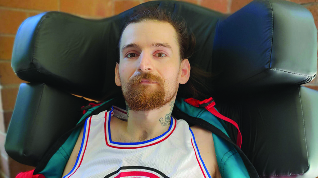 A close-up portrait of a man with a beard and neck tattoos sitting in a supportive black headrest.