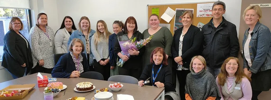 A group of colleagues gathered in an office for a celebration, with cakes and treats on a table in the foreground.