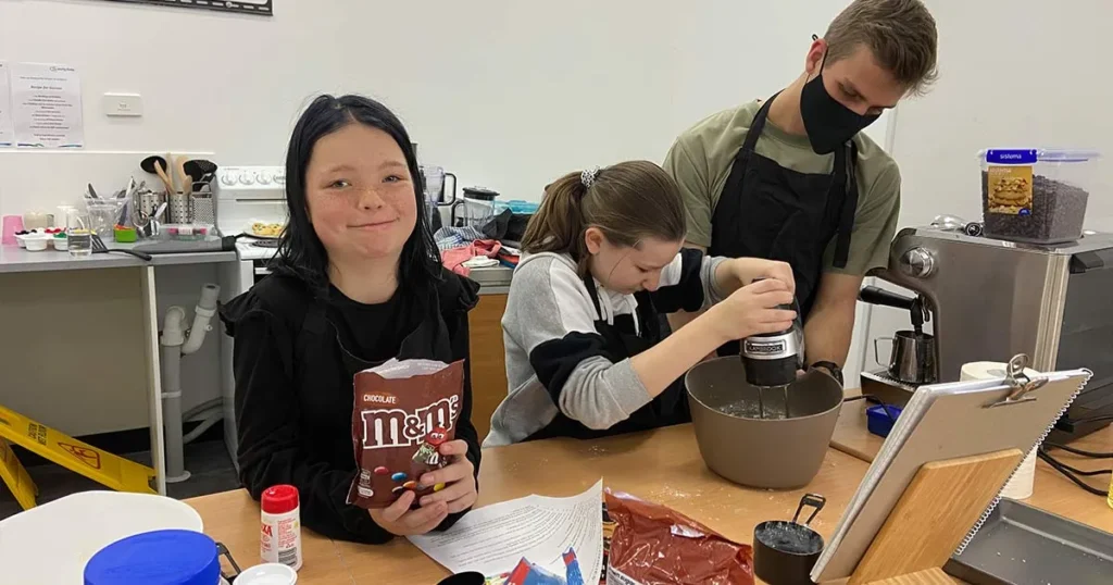 Two young girls and a young man in a mask bake together in a kitchen.