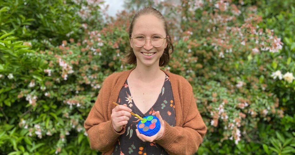 A smiling woman with glasses and an orange cardigan standing in front of green bushes while holding a small paint palette and a paintbrush.