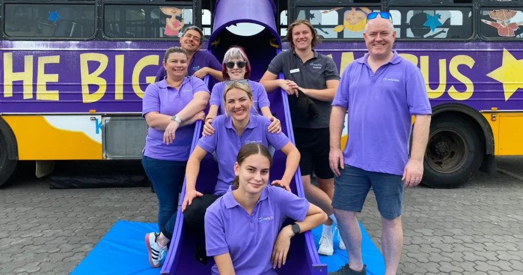 Seven people in purple "Early Links" polo shirts pose for a group photo in and around a purple slide attached to a colourful bus labelled "THE BIG BUS."