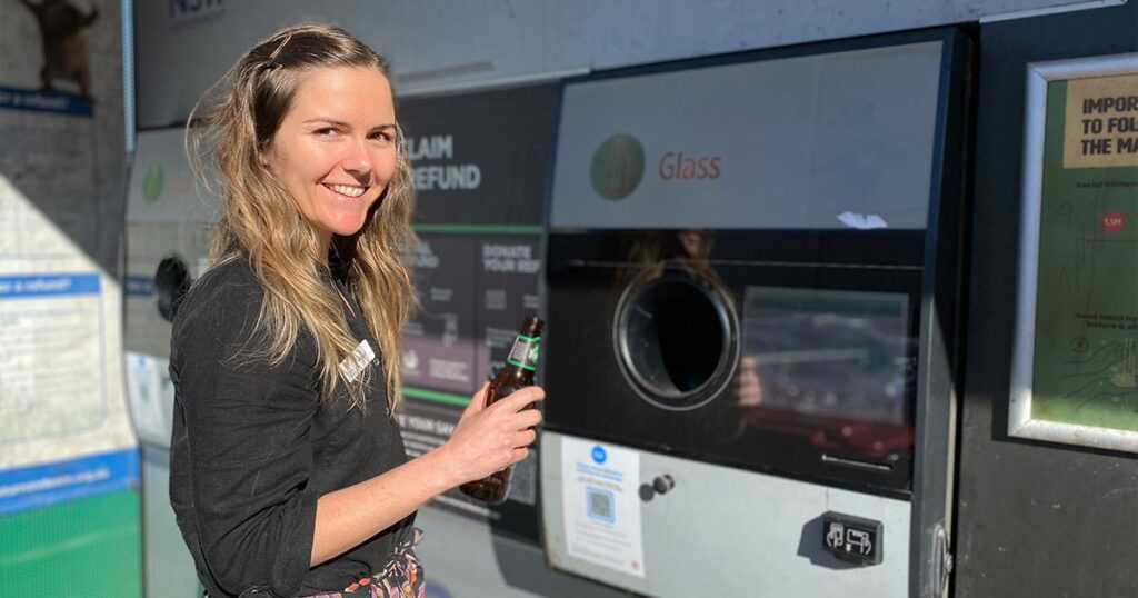 A smiling woman holding a glass beer bottle while standing in front of a "Return and Earn" reverse vending machine used for recycling.