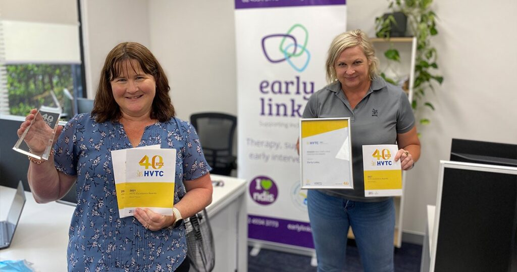 Two smiling women in an office setting holding up awards and booklets for the "40 Years HVTC Excellence Awards," standing in front of an Early Links banner.