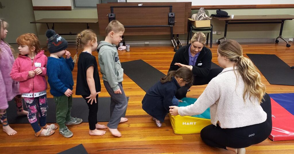 Two women supervise a group of young children in a gym or community hall, assisting a young girl as she climbs onto a yellow and blue sensory foam block on a wooden floor.