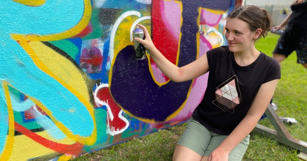 A young woman smiling as she uses a spray paint can to create a colourful mural on a large outdoor board.