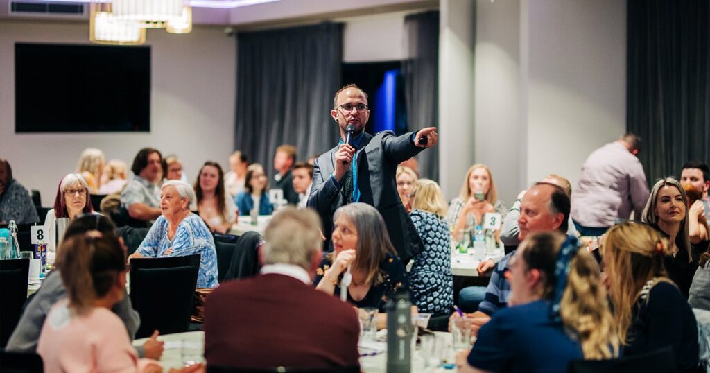 A man in a suit speaking into a microphone and pointing toward the audience during an event in a crowded banquet hall.