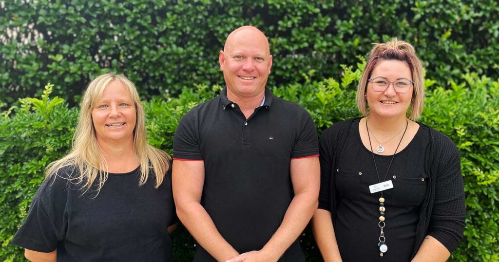 Three individuals standing side by side and smiling in front of a green hedge.