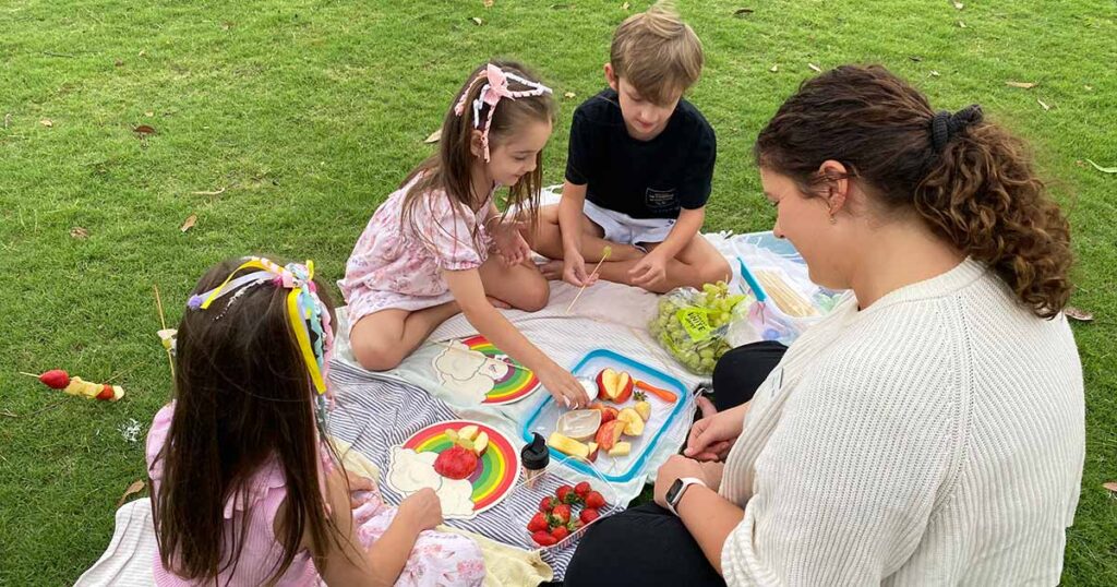 A woman and three young children enjoy a picnic on a grassy lawn