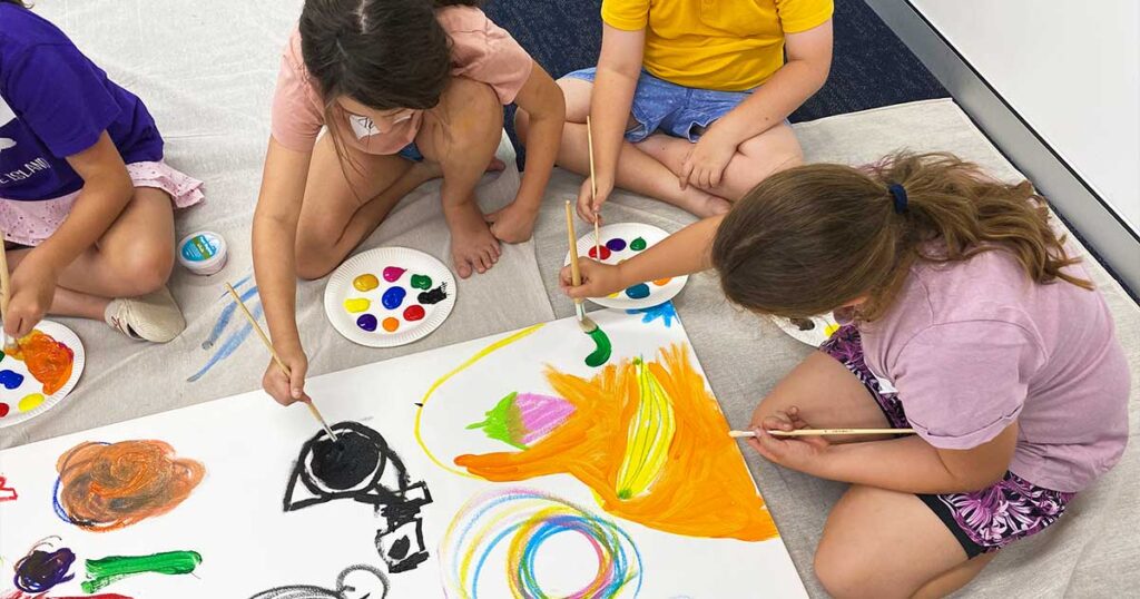 Four children sit on the floor around a large white canvas