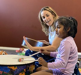 A woman holding a guitar smiles while a young girl in a purple shirt plays a colourful drum with mallets.