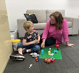 A woman in a pink shirt sits on the floor and talks with a young boy as they play with toy animals and building blocks together.