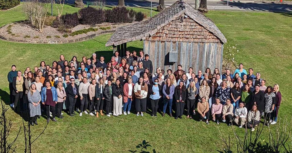 A large group of people stands together for a group photo on a grassy lawn in front of a small, rustic wooden building.