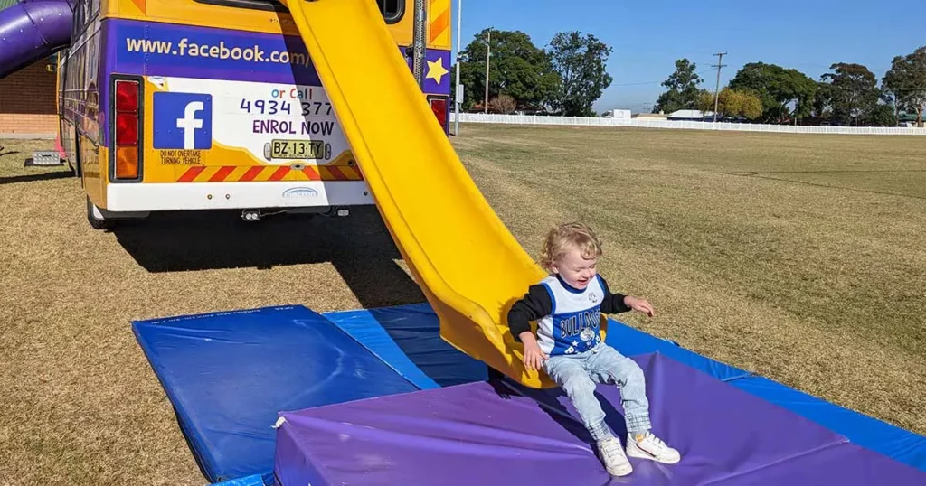 A toddler smiles while sliding down a yellow slide attached to the back of a colourful purple and yellow bus.