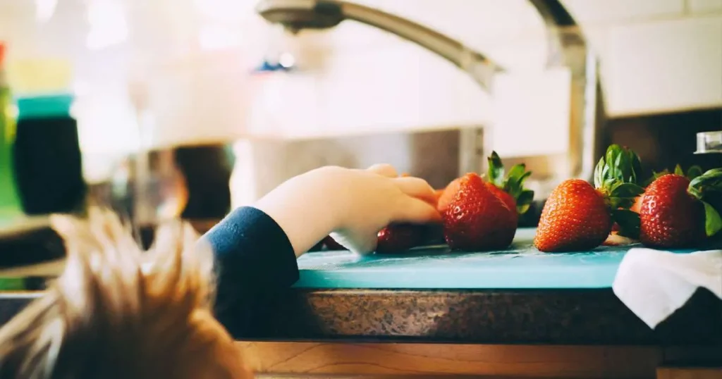 A child's hand reaches toward a group of fresh strawberries on a blue cutting board in a sunlit kitchen.