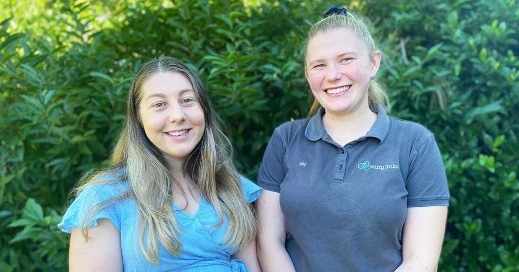 Two young women smile for a portrait against a backdrop of lush green foliage.