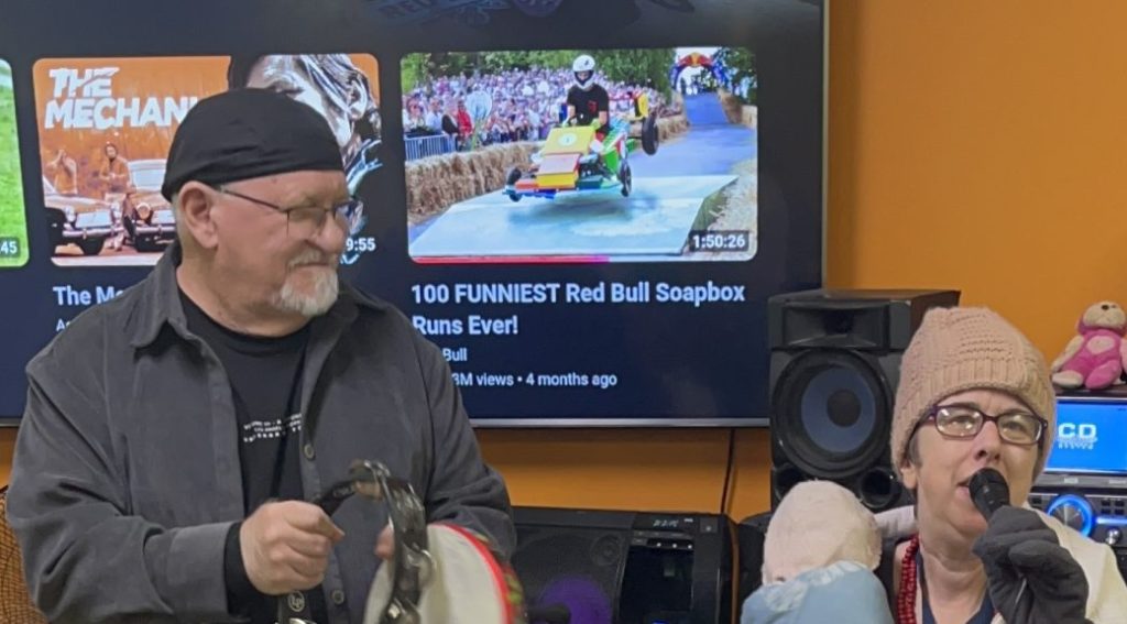 A man in a black skull cap and grey shirt playing a tambourine while standing next to a woman in a pink beanie and glasses who is singing into a microphone.