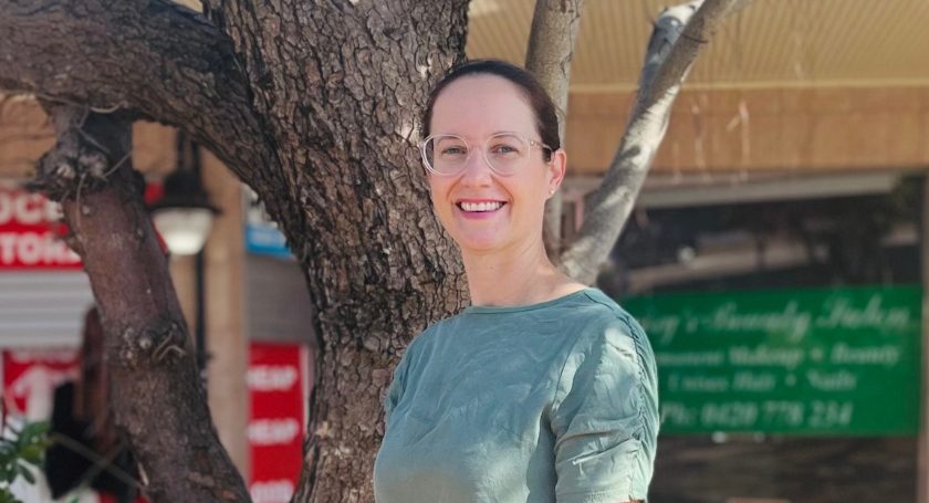 A woman with glasses and brown hair tied back, wearing a sage green blouse, smiling outdoors in front of a tree and a blurred background of shops.