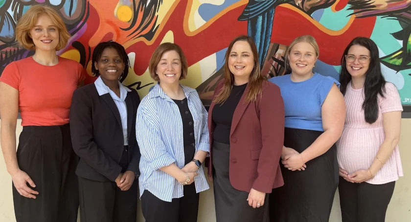 Six women of diverse backgrounds standing together and smiling in front of a colourful, abstract mural.