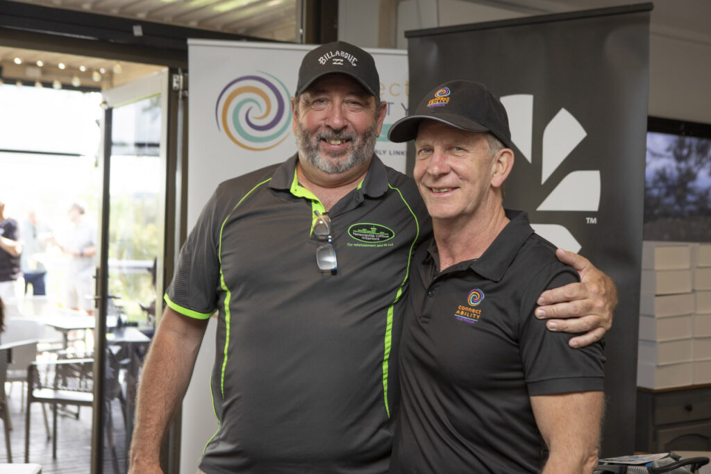 Two men in black polo shirts and caps smiling for a photo indoors